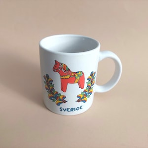 May include: White ceramic mug with a red and white Dala horse design and floral accents. The mug says "SVERIGE" at the bottom.