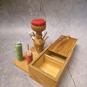 Puede incluir: Un kit de costura de madera con un cojín rojo para alfileres, soportes de hilo de madera y una caja de madera abierta. También hay carretes de hilo verde y naranja. El kit está diseñado para coser y manualidades.