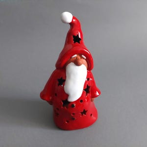 Puede incluir: Figurita de Papá Noel de cerámica roja con barba blanca y un gorro con estrellas. La figurita tiene un diseño de corte en forma de estrella y es un portavelas.