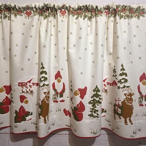 Op de afbeelding: Crèmekleurig kerstgordijn met een rode rand. Het ontwerp toont kabouters, bomen en besneeuwde landschappen. De bovenkant heeft een decoratieve rand met harten en groen, en het gordijn is versierd met sneeuwvlokken.