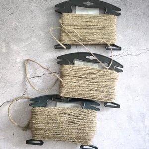 May include: Three black plastic spools each wrapped with a length of natural tan jute twine. The twine is wound horizontally around the center of each spool, with loose ends of the fibrous cord extending from the coils of the spools.