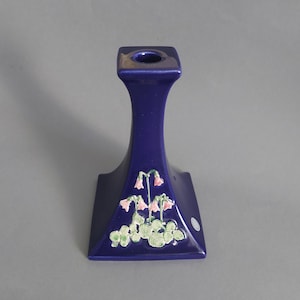 Può includere: Un portacandele in ceramica blu con una base quadrata e uno stelo alto e sottile. La base presenta un motivo floreale con fiori rosa e foglie verdi.
