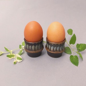 Puede incluir: Dos huevos marrones en hueveras de cerámica decorativas. Las hueveras son de color marrón oscuro con un patrón repetido de formas azules claras y blancas. Ramitas de hojas verdes están dispuestas alrededor de los huevos.