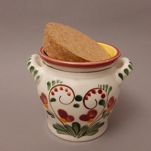 Op de afbeelding: Een witte keramische pot met een kurken deksel en een bloemenontwerp in tinten rood, groen en geel. De pot heeft twee handvatten en is versierd met een gestileerde hartvorm.