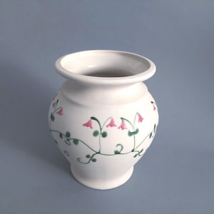 May include: A white ceramic vase with a rounded body and flared rim. The vase is decorated with pink bellflower designs and green vines. A decorative item for the home.