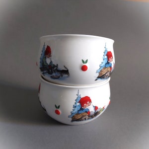 May include: Two white ceramic bowls with a Christmas theme. The bowls are stacked on top of each other. The bowls feature illustrations of gnomes in red hats and blue coats, along with trees and red berries.