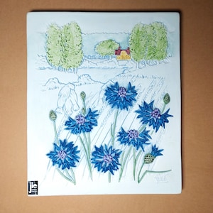 Puede incluir: Placa de pared rectangular de cerámica con un diseño en relieve de acianos azules en un campo, con una casa amarilla y árboles en el fondo. La obra de arte está firmada en la esquina inferior derecha y tiene un pequeño logotipo en la esquina inferior izquierda.