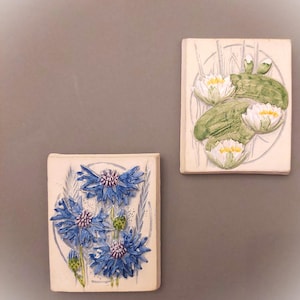 May include: Two ceramic tiles with raised floral designs. The first tile features a cluster of blue cornflowers with white centres. The second tile features white water lilies with green leaves.