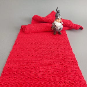 May include: A red crocheted table runner with a decorative open stitch pattern. The runner is lying flat on a grey surface with a small gnome figurine on top.