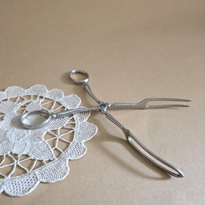 May include: Silver-toned serving tongs with a scissor-like design, featuring a ring handle and a two-pronged fork end. The tongs are resting on a white, lace doily with a scalloped edge, set against a neutral background.