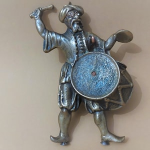 Könnte beinhalten: Eine Bronzestatue eines Mannes, der Trommel spielt. Er trägt einen Turban und hält einen Schlagstock in seiner rechten Hand. Die Trommel ist mit einem blauen und weißen Muster verziert.