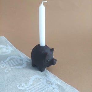 Op de afbeelding: Een zwarte keramische varkensvormige kandelaar met een witte kaars. De kandelaar heeft kleine, ronde ogen en een snuit. De kaars is wit en heeft een gebogen top. De kandelaar staat op een lichtgekleurd oppervlak.
