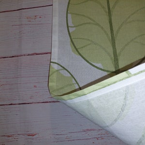 May include: White fabric with a repeating pattern of large, stylized green leaves. The leaves have a light green fill and darker green outlines. The fabric is folded, revealing a white edge and a wooden background.