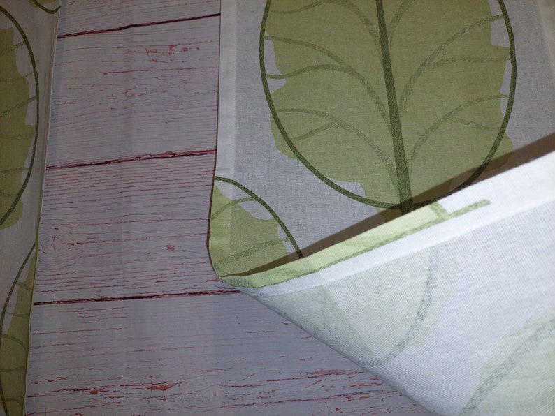 May include: White fabric with a repeating pattern of large, stylized green leaves. The leaves have a light green fill and darker green outlines. The fabric is folded, revealing a white edge and a wooden background.