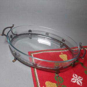 Könnte beinhalten: Eine ovale Backform aus Glas mit Metallrahmen und Griffen. Die Form steht auf einer rot-grün gemusterten Tischdecke. Der Metallrahmen hat dekorative, gebogene Griffe.