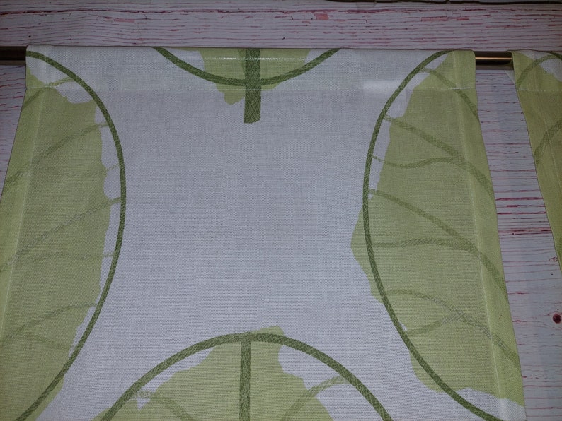 May include: Two white and green leaf-patterned curtains. The design features large, stylized green leaves with detailed vein patterns against a white background. The curtains are hanging from a gold-colored rod, adding a touch of elegance.