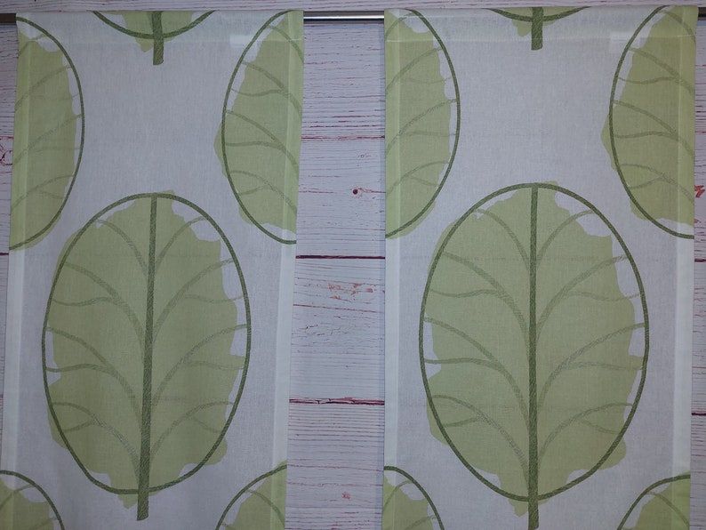 May include: Two white curtain panels with a repeating pattern of large, stylized green leaves. The leaves have a dark green outline and a lighter green fill. The curtains are hanging on a silver rod.