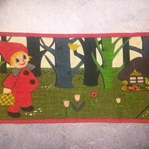 Può includere: Tessuto vintage con una scena boschiva stravagante con una figura vestita di rosso che porta un cestino. Il design include alberi, fiori e una piccola casa, il tutto incorniciato da un bordo rosso. La tavolozza dei colori è dominata da verde, rosso e nero.
