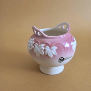 Puede incluir: Un pequeño jarrón de cerámica rosa con base y asa blancas. El jarrón está decorado con diseños de uvas y hojas blancas. Tiene una base pequeña y redonda y una abertura ancha. Probablemente es decorativo.