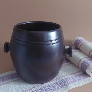 Può includere: Un contenitore in ceramica marrone scuro a forma di barile con due piccole maniglie sui lati. Il contenitore è appoggiato su un tessuto a strisce bianche e rosa.