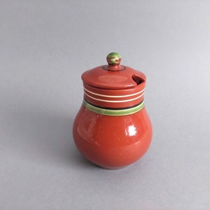 May include: A red ceramic jar with a green and white striped lid. The jar has a small spout for pouring.