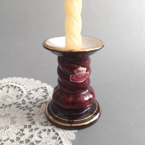 Puede incluir: Un candelero de cerámica de color rojo oscuro con un borde dorado y un interior blanco. El candelero tiene un diseño retorcido y presenta una etiqueta que dice "Bay Keramik" con una corona sobre la palabra "Bay".