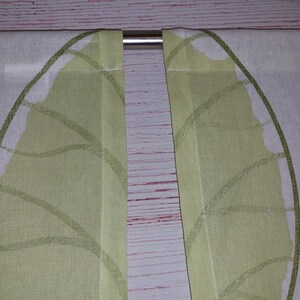May include: Two white and light green leaf-patterned curtains hanging from a silver rod. The curtains are made of a light fabric and feature a repeating leaf design in shades of green.