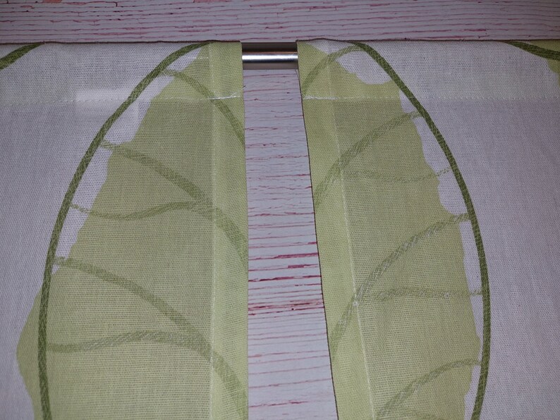 May include: Two white and light green leaf-patterned curtains hanging from a silver rod. The curtains are made of a light fabric and feature a repeating leaf design in shades of green.