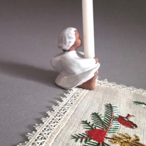 May include: A ceramic candle holder in the shape of a seated figure, holding a white candle. The figure is wearing a white robe and cap. The candle holder is on a decorative embroidered cloth with a lace edge.