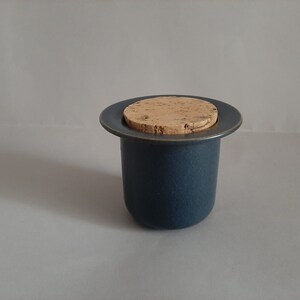 May include: A dark blue ceramic jar with a cork lid. The jar is round and has a smooth surface. The cork lid is flat and has a natural brown color.