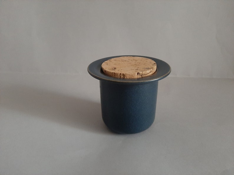 May include: A dark blue ceramic jar with a cork lid. The jar is round and has a smooth surface. The cork lid is flat and has a natural brown color.