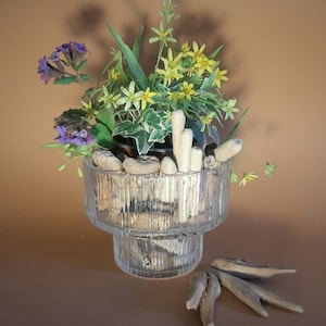 May include: A glass vase with a tiered design, filled with driftwood and a variety of yellow and purple flowers. The vase is sitting on a brown surface with additional driftwood pieces scattered around it.