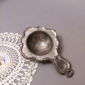 May include: A silver tea strainer with a decorative handle and a round, perforated bottom. The strainer is sitting on a white crocheted doily.