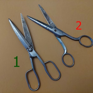 May include: Two antique metal scissors with silver blades and handles. The scissors are open, showing their sharp blades. The handles have finger holes for easy grip. The scissors are set against a brown background.
