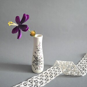 Puede incluir: Un jarrón de cerámica blanco con un diseño floral, que contiene una flor morada con estambres amarillos. Un trozo de encaje blanco de tejido abierto está drapeado cerca. El jarrón mide unos 15 cm de alto.