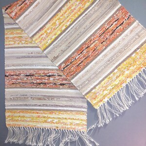 May include: A woven rug with a striped pattern in shades of beige, orange, and yellow. The rug has a fringe on both ends.