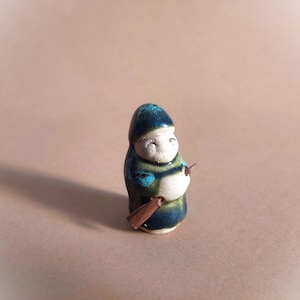 Könnte beinhalten: Eine kleine Keramikfigur einer Person, die einen blauen und grünen Hut und eine Robe trägt und einen braunen Stock in der rechten Hand hält. Die Figur hat ein weißes Gesicht mit einem lächelnden Ausdruck.