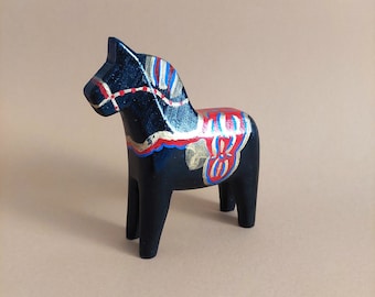 May include: A black wooden horse figurine with red, blue, and gold painted details. The horse has a saddle and bridle, and is a traditional Swedish Dala horse.
