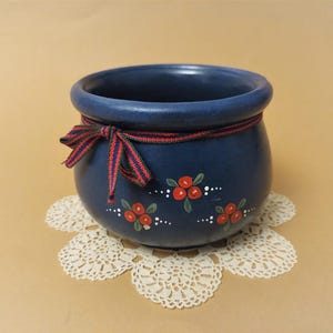 May include: A navy blue ceramic pot with a decorative red and white ribbon tied around the rim. The pot features hand-painted red berries and white dots. It rests on a white lace doily.