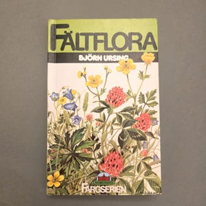 May include: A vintage book titled "Fältflora" by Björn Ursing, with a cover illustration of wildflowers. The cover has a green header with white text, and the book showcases various flowers in shades of yellow, red, and blue.