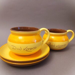 Könnte beinhalten: Zwei gelbe Kaffeetassen aus Keramik mit braunen Rändern und Henkeln. Die Tassen haben ein gelbes und weißes Streifenmuster und den Text "Din Kopp" und "Min Kopp" in Braun auf der Vorderseite.