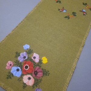 May include: Olive green table runner with a fringed edge. The runner is decorated with painted flowers in shades of red, pink, purple, blue, and yellow. Small birds are also painted on the runner.