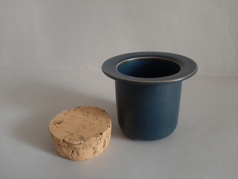 May include: A dark blue ceramic container with a cork lid. The container has a wide rim and a smooth, rounded bottom.