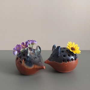 May include: Two ceramic animal-shaped vases with holes for flowers. The vase on the left is blue and brown with purple flowers. The vase on the right is blue and brown with a yellow flower.