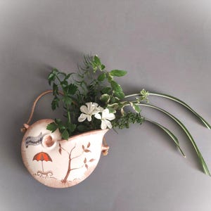 May include: A white ceramic wall planter shaped like a seashell with a painted design of a tree and an umbrella. The planter is filled with green plants and white flowers.