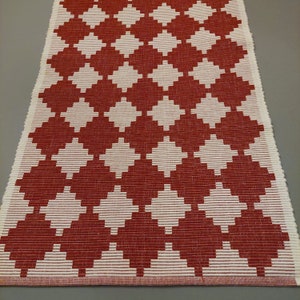 Op de afbeelding: Een rood en wit geweven tapijt met een geometrisch ruitpatroon. Het tapijt heeft een gefranst randje.