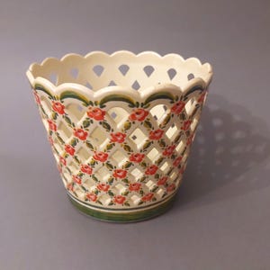 May include: A white ceramic planter with a lattice design and red and green floral pattern. The planter has a scalloped rim.
