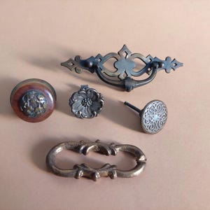 Può includere: Cinque pomelli e maniglie per armadi di stile antico in vari design. I pomelli sono in metallo e hanno una finitura marrone scuro. Un pomello ha un design floreale, un altro ha un design circolare e il terzo ha un design rotondo con un centro rialzato. Le maniglie sono anch'esse in metallo e hanno una finitura marrone scuro. Una maniglia ha un design semplice e curvo, mentre l'altra ha un design più elaborato con un motivo floreale.