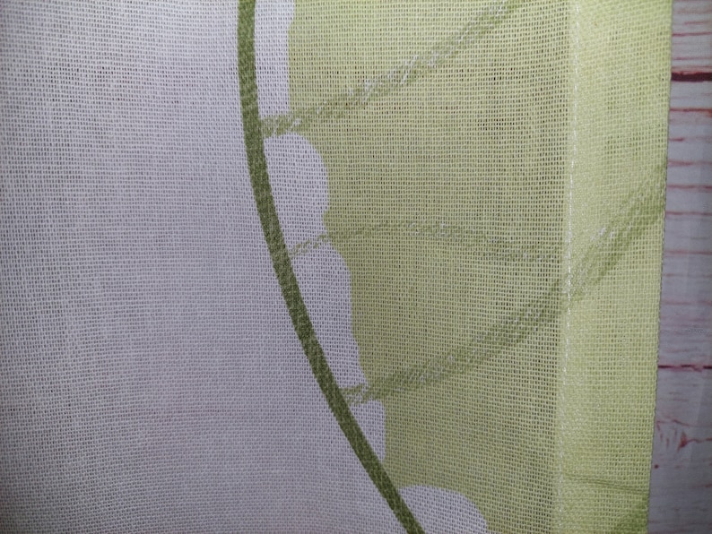 May include: Close-up of a sheer fabric with a floral design. The fabric features a white background with a green leaf pattern and a solid light green border. The fabric is likely used for curtains or home decor.