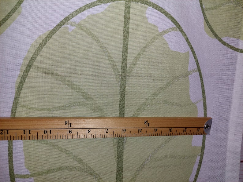 May include: Close-up of fabric with a large green leaf pattern on a white background. A wooden ruler shows measurements in inches. The fabric has a light green leaf design with darker green outlines.